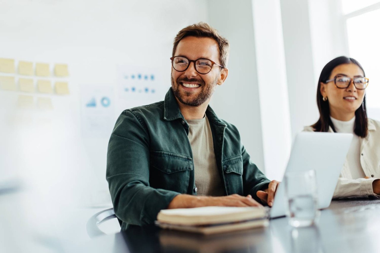 A smiling male coworker with a laptop sitting next to a female coworker with glasses.