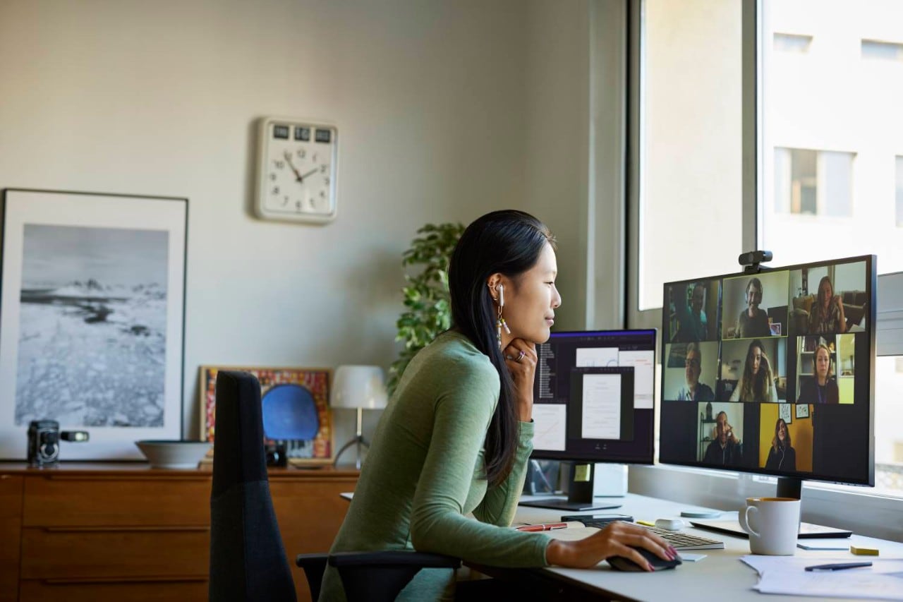 A woman sitting in an office, wearing earphones and actively participating in a live video conference call with multiple people.